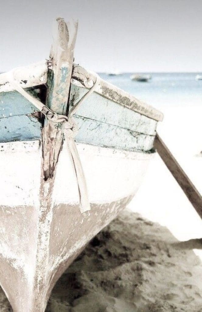 Travel inspired image of traditional wooden fishing boat with aged patina of light turquoise blue and white stripes pulled up onto soft white sand