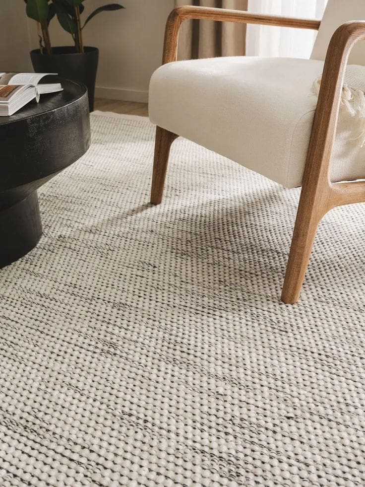 Woven loop rug in warm neutral off-white and light grey fleck, with wooden frame armchair and rounded coffee table in dark mahogany brown