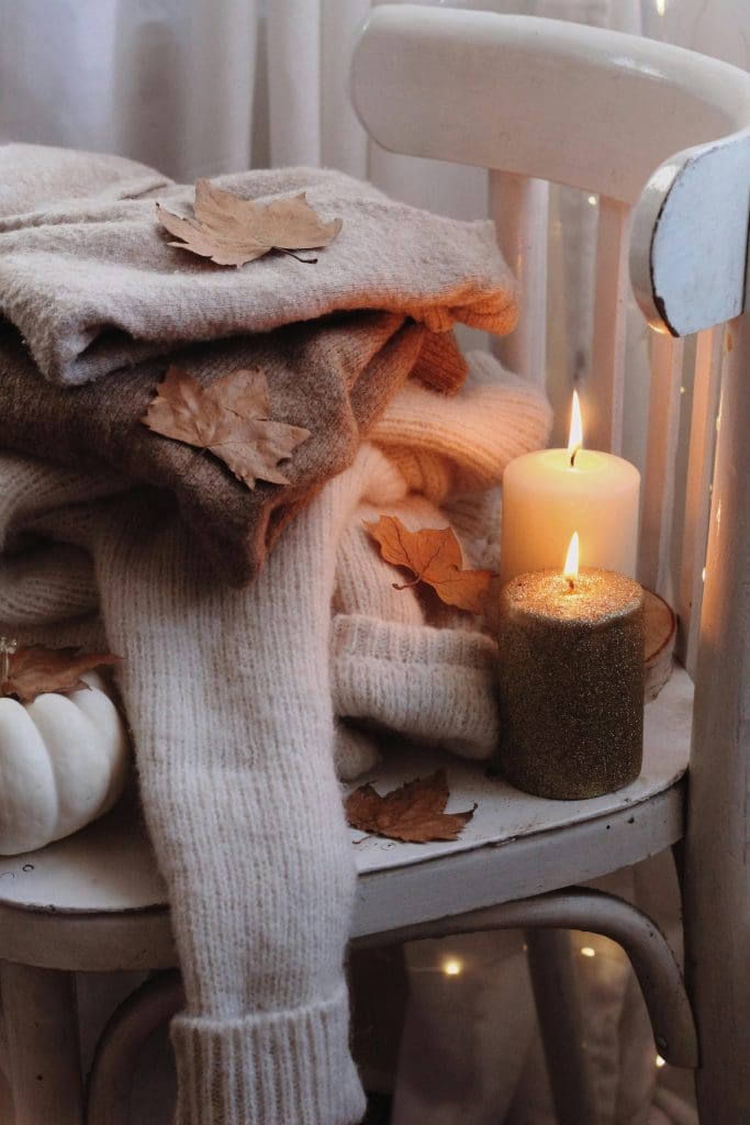 Worn patina dining chair piled with warm and cozy warm white jumpers, autumnal leaves of oranges and browns as well as 2 lit pillar candles