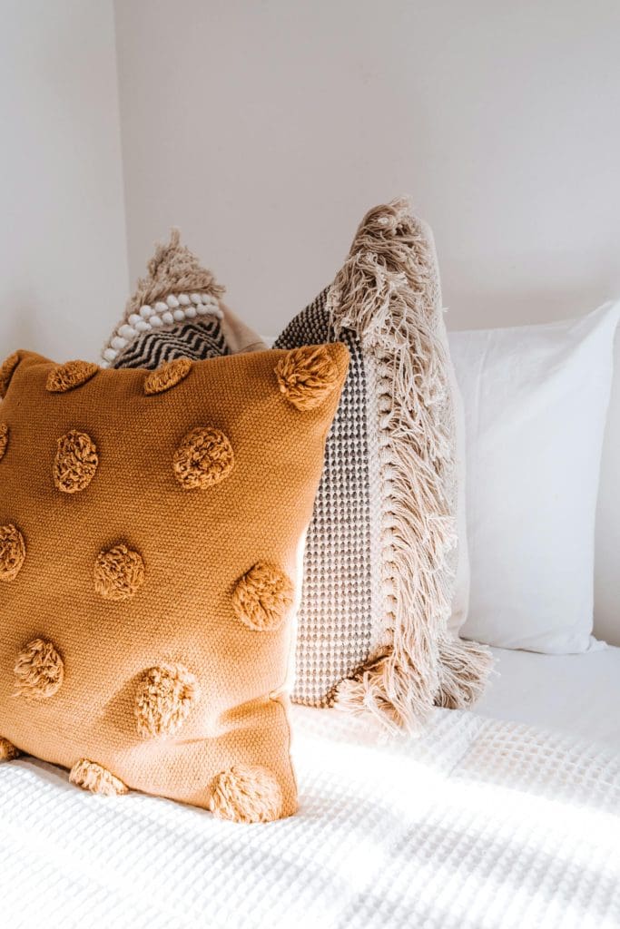 Biophilic Interior Design image of white waffle bedding and decorative throw cushions, hand woven jute and fringed cushion with camel coloured pom pom cushion in front. Picture filled with natural light