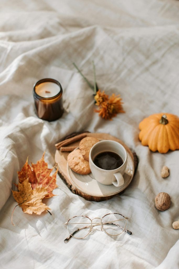 Off-white cheesecloth bed scattered with Autumn leaves in shades of orange and yellow with a cup of coffee and biscuits on an off-cut of wood