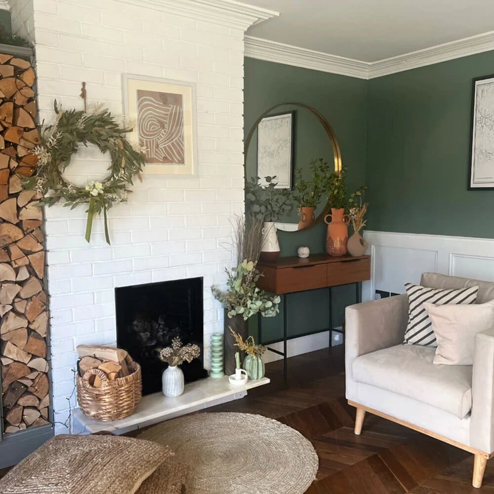 Lounge with forest green wall, white brick feature fireplace and styled with rustic chopped wood, faux foliage and stems and a large round brass framed mirror