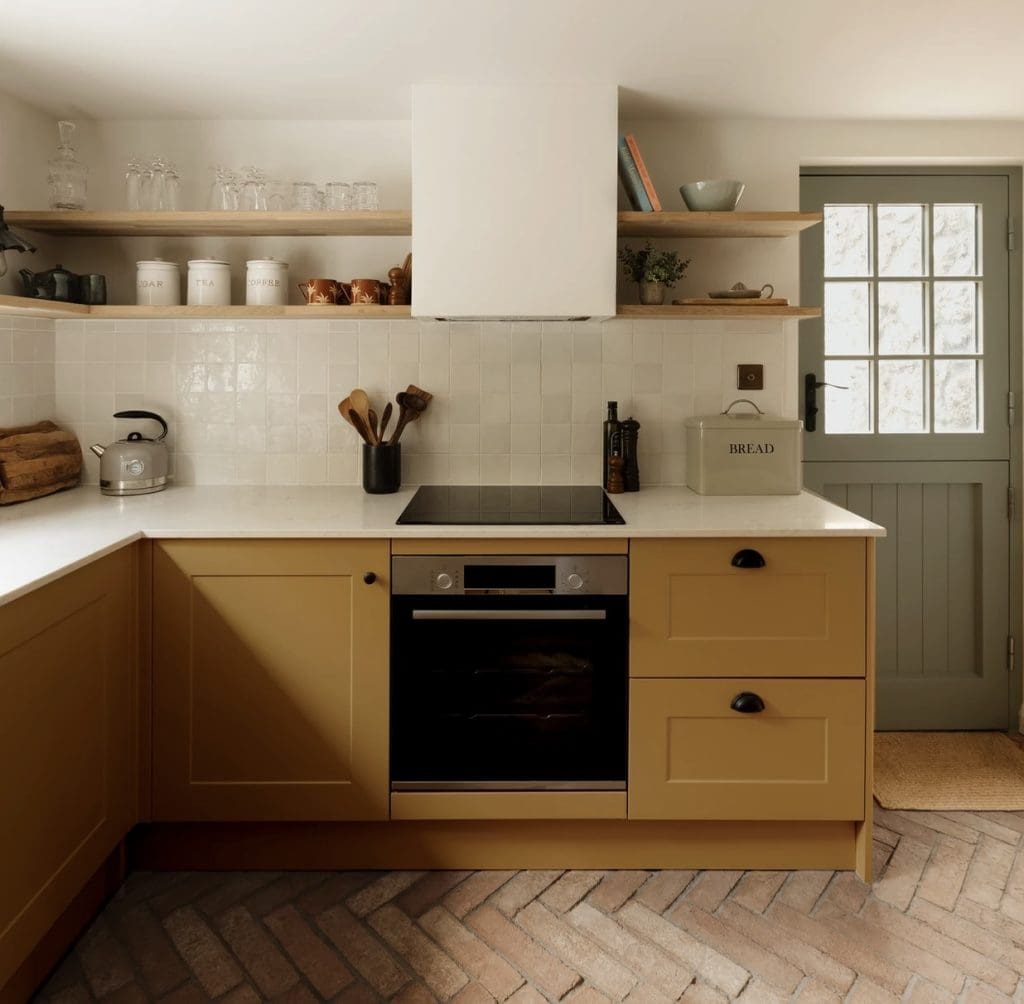 Yellow ochre kitchen lower cupboards with glazed white tile splashback and open shelving styled with glassware, white storage canisters and books.