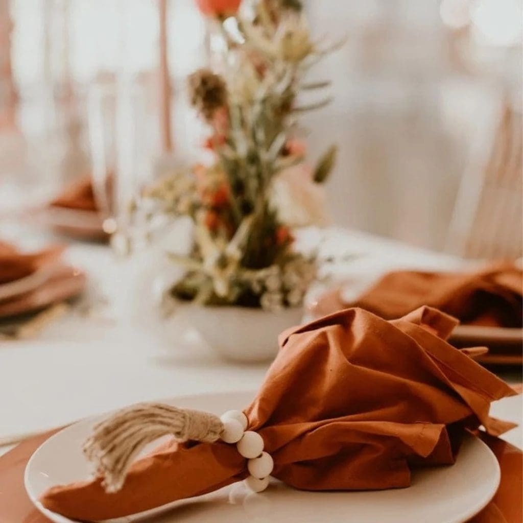 Tablescape with white cloth, light terracotta tapered candles and dark terracotta cloth napkins with wooden bead napkin rings