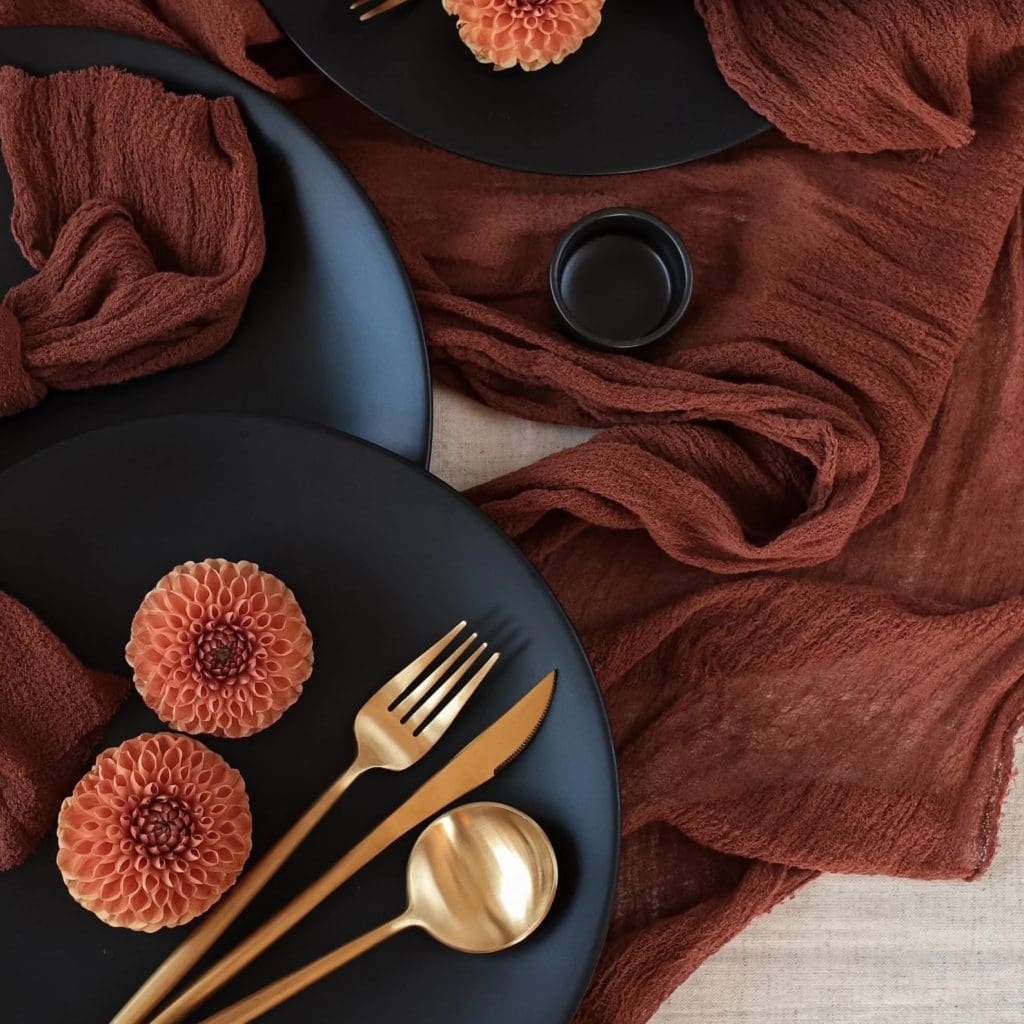 Dark and moody tablescape with terracotta cheesecloth table runner, black plates and gold cutlery