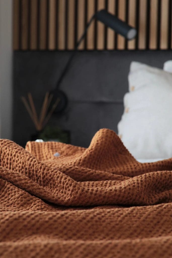 Waffle fabric throw in orange-terracotta on bed with white pillows, dark chocolate upholstered headboard and wooden slatted wall detail