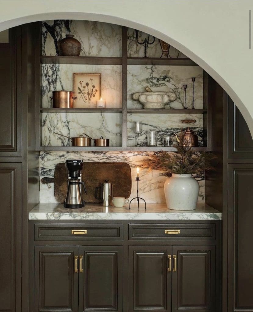 Marble arched in-built coffee nook, featuring dark forest green cabinetry, vintage wooden boards and large white ceramic vase with foliage.