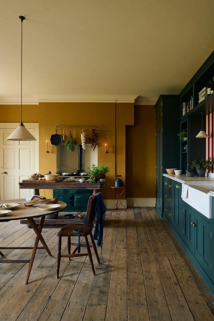 A dark golden mustard painted wall in a dark teal kitchen space. Reclaimed hardwood flooring and low hung pendant lighting